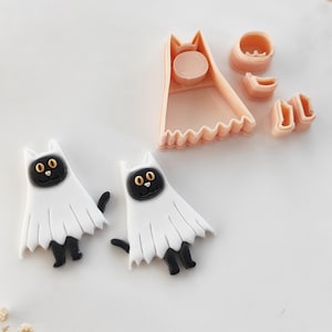Könnte beinhalten: Eine Sammlung von Halloween-Themen-Katzen-Ausstechern. Zwei weiße katzenförmige Figuren mit schwarzen Beinen und Schwänzen befinden sich im Vordergrund. Die Katzen tragen weiße Geisterkostüme mit schwarzen Augen und Nasen. Zusätzliche Ausstecherteile sind im Hintergrund.