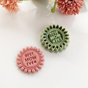 Könnte beinhalten: Zwei sonnenblumenförmige Ausstecher. Einer ist rosa mit dem Text "BEST MOM EVER", und der andere ist grün mit dem Text "MOM 83V3". Die Ausstecher befinden sich auf einer weißen Oberfläche mit rosa Blumen im Hintergrund.