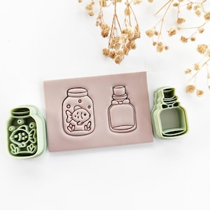May include: Green and pink cookie cutters shaped like jars, one with a fish design. A pink rectangular stamp with jar designs is also present. These cutters are for making biscuits or other treats.