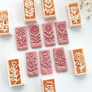 Op de afbeelding: Een verzameling bloemvormige koekjesstempels. De stempels zijn rechthoekig, met een roze basis en een beige frame. Elke stempel heeft een ander bloemontwerp, waaronder madeliefjes, zonnebloemen en andere botanische illustraties.