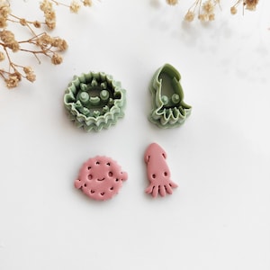 May include: Four colorful cookie cutters in the shapes of sea creatures. Two are green, one is a round shape with a face and the other is a squid. The other two are pink, one is a pufferfish and the other is a squid.
