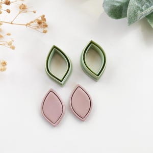 May include: Four leaf-shaped polymer clay cutters and two leaf-shaped clay pieces. The cutters are light green, and the clay pieces are a muted pink. The cutters are nested, creating a layered effect. The clay pieces are solid.