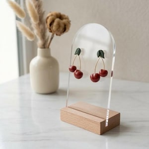 Op de afbeelding: Kersen oorbellen met rode vruchten en groene bladeren, tentoongesteld op een transparante acryl standaard met een houten voet. Op de achtergrond staat een vaas met gedroogde bloemen.