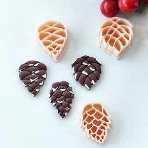 May include: An assortment of pine cone-shaped cookie cutters and decorative pine cones. The cutters are light beige, while the pine cones are brown with white accents. Red berries are visible in the background. Ideal for baking and crafting projects.