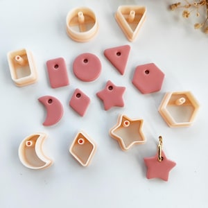 Könnte beinhalten: Eine Sammlung von rosafarbenen und beigefarbenen Polymer Clay Ausstechern in verschiedenen Formen, darunter Kreise, Dreiecke, Sterne und Sechsecke. Einige Ausstecher haben ein Loch zum Aufhängen. Die Formen sind auf einer weißen Oberfläche angeordnet.