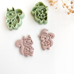 May include: Two light green plastic cookie cutters and two corresponding light pink clay shapes. One cutter and shape depict a squirrel wrapped like a mummy, while the other depicts a squirrel wearing a witch hat and holding a small acorn.