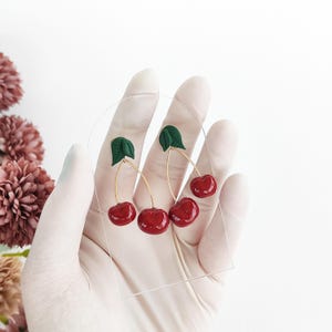 Op de afbeelding: Kersen oorbellen. Elke oorbel heeft twee glanzende rode kersen met groene bladeren, die aan goudkleurige draden hangen. De oorbellen worden tegen een witte achtergrond getoond.