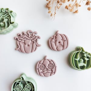 May include: Three mauve-coloured pumpkin-shaped cookie stamps featuring distinct facial designs, including a Medusa-inspired pumpkin with snakes, a sad pumpkin, and a pumpkin with a moustache and gear-shaped eyes, alongside two green plastic cutters.