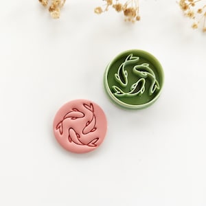 May include: A green and white plastic cookie cutter featuring three koi fish designs. A pink cookie with the same koi fish design is next to the cutter. The koi fish are arranged in a circular pattern.