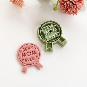 Może przedstawiać: Dwa wykrawacze do ciastek w kształcie wstążki. Jeden jest różowy i mówi "BEST MOM EVER" z sercem. Drugi jest zielony i mówi "MOM BEST" z sercem. Oba mają ogony wstążki.
