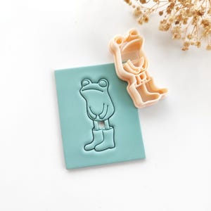 Può includere: Un quadrato azzurro con un'impronta a forma di rana e un tagliabiscotti a forma di rana abbinato. La rana indossa stivali e ha un'espressione sorridente. Il tagliabiscotti è di colore pesca chiaro.