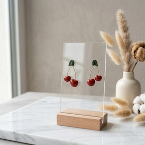 Op de afbeelding: Een paar kersen oorbellen met rode kersen, groene bladeren en goudkleurige stelen, gepresenteerd op een transparante acryl standaard met een houten voet. Op de achtergrond een vaas met droogbloemen.