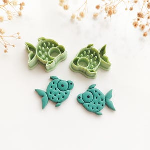 May include: Two light green plastic fish-shaped cookie cutters and two corresponding teal-colored fish-shaped cutouts made of clay or dough. The cutters feature internal details for scales and eyes, while the cutouts show the embossed texture.