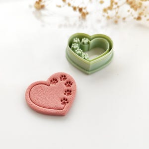 Peut inclure: Un emporte-pièce en forme de cœur et un biscuit rose avec des empreintes de pattes. L'emporte-pièce en plastique vert a une forme de cœur avec des empreintes de pattes à l'intérieur. Le biscuit rose a une forme de cœur avec des empreintes de pattes sur le côté. Le fond est blanc.