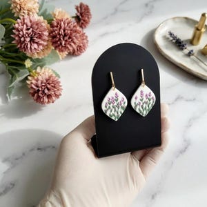 May include: A pair of white, diamond-shaped earrings with gold-coloured posts. Each earring has a lavender floral design with green stems. The earrings are displayed on a black stand, with flowers in the background.