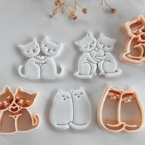 May include: A selection of cat-shaped cookie cutters in different designs. The cutters are white and beige, depicting cats hugging, holding hearts, and cuddling. These detailed designs are ideal for baking themed biscuits.