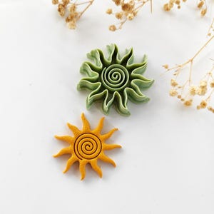 Pode incluir: Dois cortadores de biscoitos em forma de sol. Um é de cor amarelo-alaranjado brilhante com um centro em espiral e o outro é verde claro. Os cortadores têm uma borda ondulada e são provavelmente usados para assar ou fazer artesanato.
