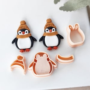May include: A selection of penguin-shaped cookie cutters and two penguin figurines. The penguins are black, white, and red, wearing brown hats and scarves. The cutters are light pink and include hat, scarf, and penguin shapes.