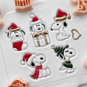 May include: A collection of white Snoopy character designs in festive Christmas attire. Each Snoopy figure is wearing a Santa hat, scarf, or holding a Christmas tree. Some are paired with gift boxes and stockings. The designs are accented with red, gold, and green.