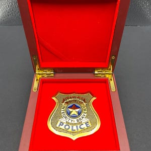 May include: A gold-colored police badge with a blue and white star emblem, displayed inside a red velvet-lined wooden box. The badge reads "Officer, Integrity, Trust, Bravery, Police". The box is open, revealing the badge.