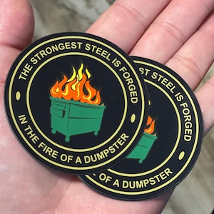 May include: Two black and gold round stickers with the text "The strongest steel is forged in the fire of a dumpster". The stickers feature a green dumpster with flames coming out of it.