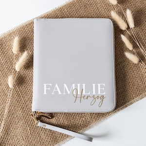 Könnte beinhalten: Eine graue Reißverschlusstasche mit der Aufschrift "FAMILIE Herzog" in Weiß. Die Tasche hat einen goldenen Reißverschluss und einen grauen Riemen.