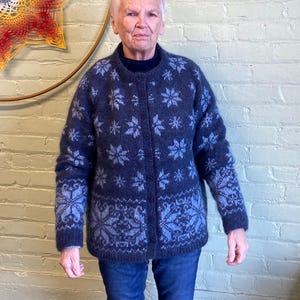 Op de afbeelding: Een marineblauwe cardigan met een sneeuwvlokpatroon in lichtblauw. De trui met knopen heeft lange mouwen. De persoon op de foto draagt een blauwe spijkerbroek.