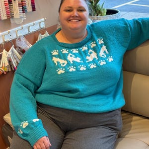 May include: A teal sweater with white dog and paw print designs. The sweater has a crew neck and long sleeves. The cuffs of the sleeves also feature the paw print design. The person in the image is wearing gray sweatpants.