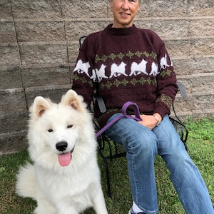 May include: A woman wearing a maroon sweater with a white dog pattern sits in a black metal chair with a white dog sitting in front of her. The woman is wearing blue jeans and blue sneakers. The dog has a purple leash.