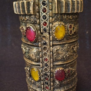 Può includere: Un bracciale a polsino antico in ottone con intricati motivi in rilievo e pietre preziose colorate. Il bracciale presenta pietre rosse, gialle e verdi ed è progettato con più fasce e una chiusura a cerniera. Il metallo ha un aspetto invecchiato.
