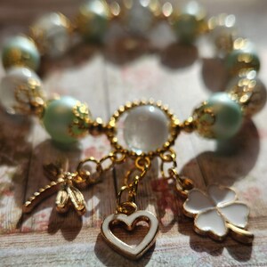 May include: A delicate bracelet with green and white beads, a gold charm with a clear crystal, and three dangling charms: a heart, a four-leaf clover, and a dragonfly.