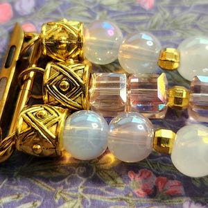 May include: Gold-toned Apple Watch band with a mix of clear, iridescent, and white beads. The band features decorative gold accents and a secure clasp. The beads are round and faceted, creating a stylish accessory.
