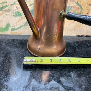 May include: A copper coffee pot with a black wooden handle and a brass spout. The pot is sitting on a black surface with a measuring tape next to it. The tape reads 3 inches.