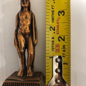 May include: A bronze-colored figurine of a Native American man standing on a rectangular base. The figurine is approximately 4 inches tall.