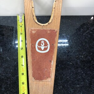 May include: Wooden boot jack with a brown leather pad and a white logo with the letter "S" inside a circle. The boot jack is 11 inches tall.