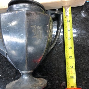 May include: A black metal teapot with a silver-colored inscription of the letter "G" on the side. The teapot has a round base and a handle. A yellow measuring tape is visible in the background.