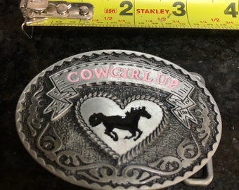 May include: Silver-toned oval belt buckle with the words "COWGIRL UP" in pink. The buckle features a heart with a black horse silhouette. The tape measure shows the buckle is approximately 3 inches long.