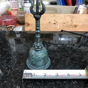 May include: A detailed, antique-style bell with a trident-shaped top. The bell has a weathered, teal-green finish and a textured surface. The bell is approximately 5 inches tall.