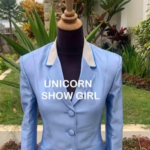 Op de afbeelding: Een lichtblauwe blazer met een witte kraag en een blauwe bies. De blazer heeft drie knopen en twee zakken. De tekst "UNICORN SHOW GIRL" is op de voorkant van de blazer gedrukt.