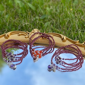 May include: Three layered beaded bracelets with different coloured stones. The bracelets are made of dark red beads and feature a silver charm with a purple stone at the end. One bracelet has a silver charm with an orange stone.
