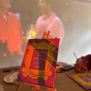 May include: A pink canvas with a yellow frame painted on it. The frame has the words "I'll be there for you" written inside. The canvas is on a wooden easel.
