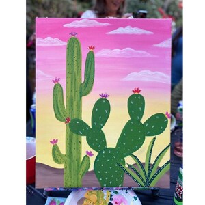 May include: A hand-painted canvas featuring a desert scene with green cacti and pink flowers. The background transitions from a yellow-orange to a pink sky with white clouds. The painting is on a wooden easel.