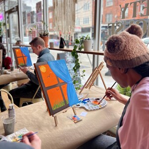May include: People painting at a workshop. Each person is working on a small canvas depicting a window scene with an orange sky, blue curtains, and a yellow sun. The canvases are on small wooden easels.