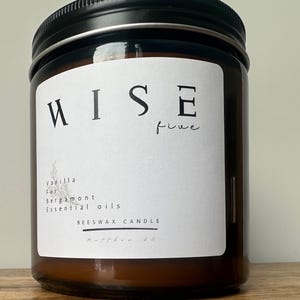 Puede incluir: Una vela en tarro de cristal marrón con tapa negra. La etiqueta dice "RISE five" con "Vanilla, Fur, Bergamot, Essential oils, Beeswax Candle" impreso debajo. La vela está sobre una superficie de madera.