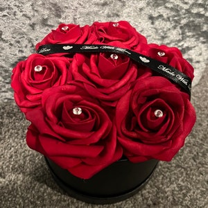 May include: A black box filled with red roses, decorated with a black ribbon and rhinestones. The ribbon has the text "Made With Love" on it.