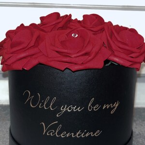 May include: A black gift box with a red velvet rose arrangement. The box has the words "Will you be my Valentine" written in gold lettering.