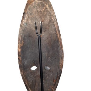 May include: A wooden mask with two holes for eyes and a metal rod attached to the back. The mask is dark brown and has a rough texture.