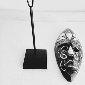 May include: Black and white wooden mask with intricate carvings and a stand. The mask has a human face with a wide mouth and large eyes.