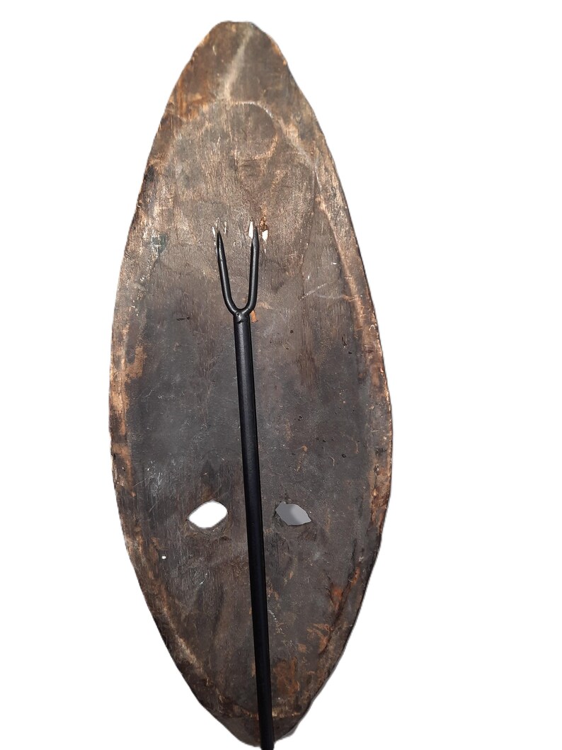 May include: A wooden mask with two holes for eyes and a metal rod attached to the back. The mask is dark brown and has a rough texture.