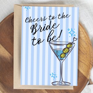 Pode incluir: Um cartão com fundo listrado azul e branco e o texto "Cheers to the Bride to be!". O cartão apresenta uma ilustração de copo de martini com azeitonas. O cartão está dentro de um envelope marrom.